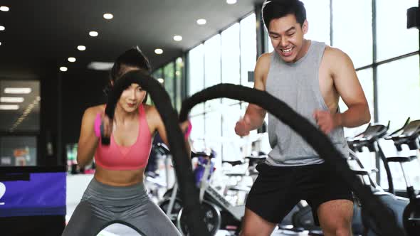 Asian Personal Trainer Coaching a Bodybuilding Woman to Perform Battle Ropes Exercise in Fitness Gym