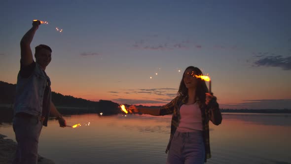 Young Happy Multiethnic Couple Holding Burning Sparkling Candles and Running By the Sea During