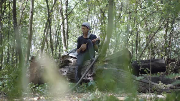 Extreme Wide Shot of Young Man Sitting on Log in Forest and Playing Ukulele. Portrait of Sad Male