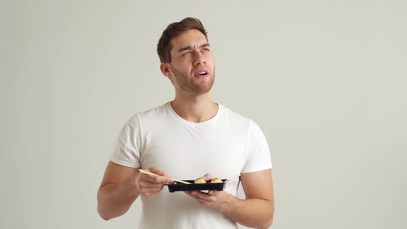 Studio Portrait of Displeased Young Man Eating Unappetizing Sushi Rolls with Chopsticks on White