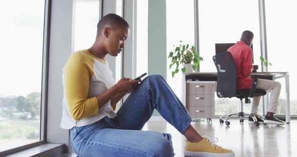 African american creative businesswoman sitting on floor using smartphone, male colleague at desk