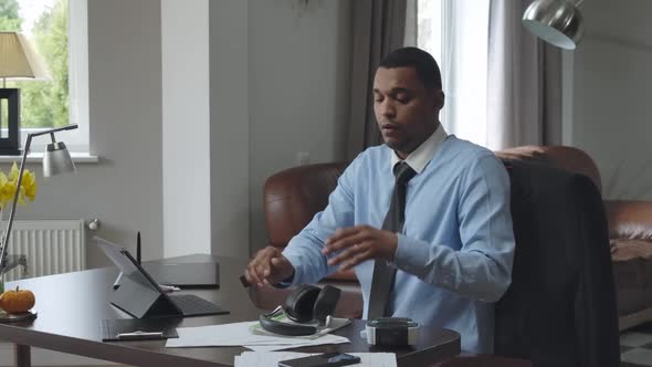 Tired African American Man Loosing Necktie Taking Off Headphones After Virtual Conference in Home