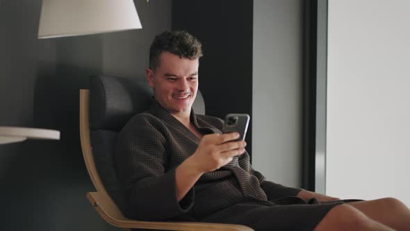 Happy Relaxed Man is Chatting Online By Smartphone Sending Message and Smiling Sitting in Lounger