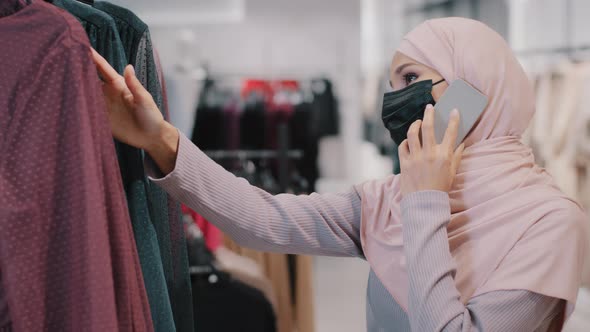 Young Arab Woman in Hijab Wearing Medical Mask Chooses Dress in Clothing Store Speaks on Phone