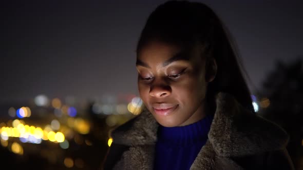 A Young Black Woman Works on a Device with a Smile Then Smiles at Camera at Night  Face Closeup