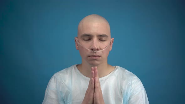 Bald Young Man with Oncology Looks at the Camera and Prays on a Blue Background. The Patient Folded
