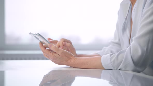 Old woman in white blouse wrinkly hands type on smartphone