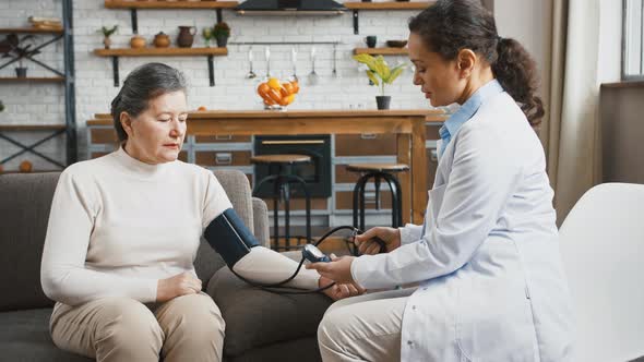 Woman Doctor Measuring Blood Pressure of Elderly Female with Bloodpressure Cuff Sitting on Couch