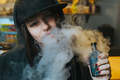 Young pretty woman in cap smoke an electronic cigarette at the vape shop. Hip-hop style. Closeup - PhotoDune Item for Sale