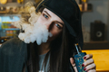 Young pretty woman in cap smoke an electronic cigarette at the vape shop. Hip-hop style. Closeup - PhotoDune Item for Sale