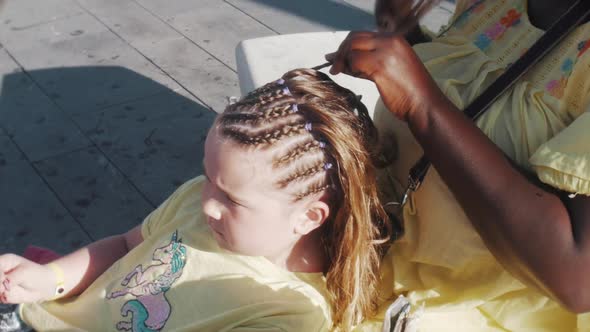A young girl has her hair braided in plats on the street sidewalk. The hairdresser lady platting and