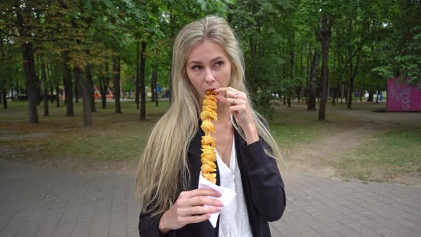 A Young Beautiful Girl with Blond Hair in a White Dress and a Black Cardigan Eats a Street Food