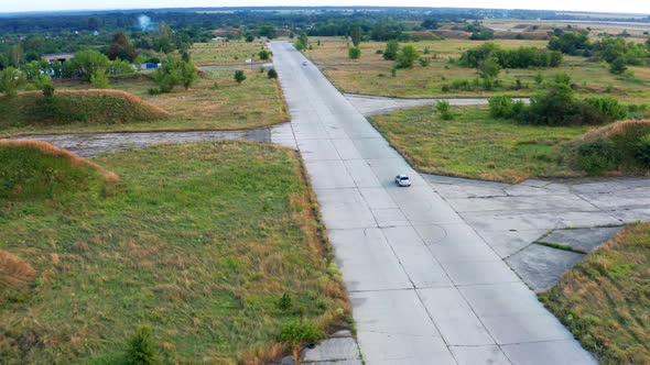 Flying over a car that is traveling on an airplane runway. Forest and grass on the sides.