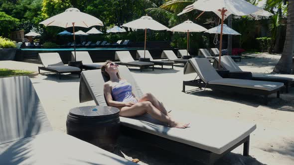 Young Sexy Woman in Swimsuit Wearing Sunglasses Resting Alone Lying on Lounge Chair on Beach During