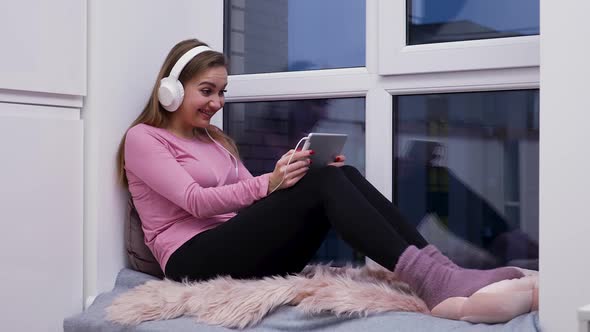 Young Woman Sits on Windowsill Near Large Window Wearing Big White Headphones Watching Movie on