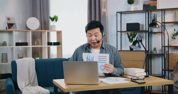 Asian Man in Headset Sitting in Home Office Near Laptop and Holding Online Meeting