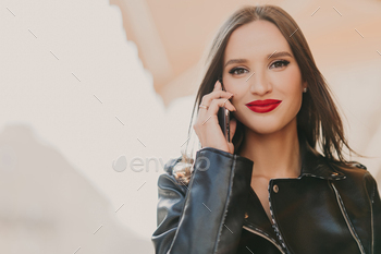, talks on mobile phone with best friend, dressed in fashionable clothes, wears red lipstick, makeup, stands on blurred background of some building