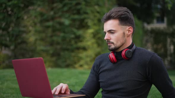 Medium Shot of Positive Successful Young Man Surfing Internet on Laptop Laughing Sitting in Park