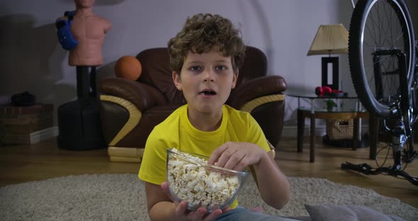 Portrait of Little Caucasian Boy Watching TV and Chewing Popcorn. Cute Curly-haired Teenage Kid