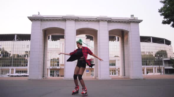 Young Stylish Funky Girl with Green Hair Riding Roller Skates and Dancing in Park