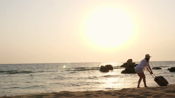 Young Woman in Short Shorts, Sun Hat and Sunglasses, Dragging a Suitcase Along the Beach By the Sea