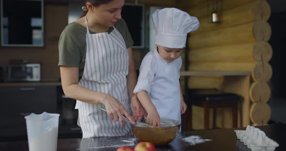 Mom and Son in Chef's Uniform Knead the Dough with Whisk in the Kitchen