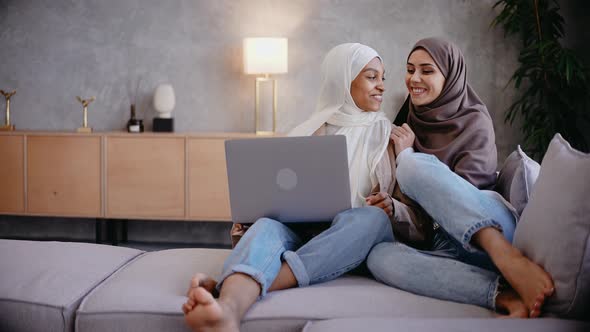 Muslim Girl Sits on Couch with Her Friend Looking at the Laptop in Bright Studio