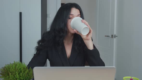 Woman Is Using Laptop While Sitting at Her Desk and Drinking Coffee. Young European Businesswoman Is