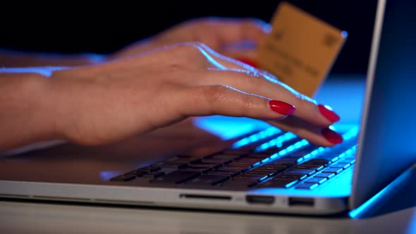 A Woman Uses a Laptop to Shop and Pay Online with a Credit Card in Office at Home in Blue Neon Light