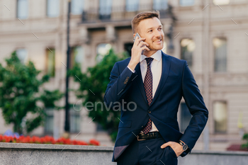 e prosperous young male entrepreneur makes phone call in roaming, uses tariffs, stands against city blurred background, discusses something.