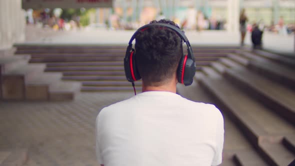 A Young Handsome Man in a White Tshirt and Headphones Sits on the Steps in the City and Listens to