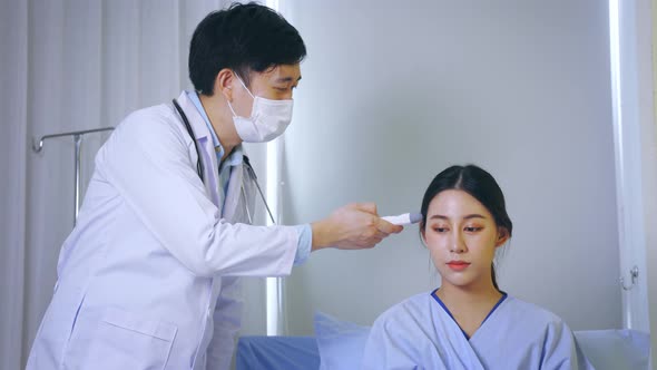 Young Male Doctor Examining Asian Female Patient Using Thermometer for Temperature Check in Hospital