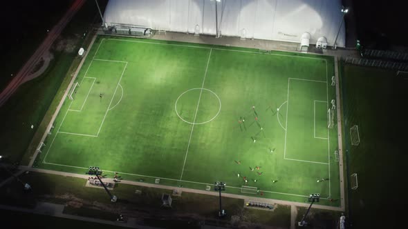 Aerial view: Players players play soccer game at night on a green field.