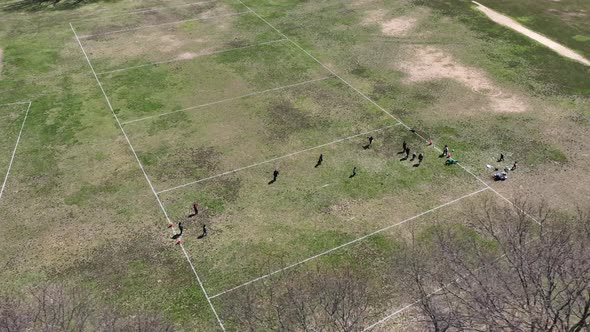 An aerial view over a park next to a parkway. People are playing soccer on a dry field on a sunny da