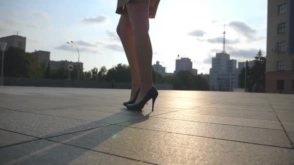 Slim Female Legs in Black Shoes on High Heels Walking on City Square at Sunset. Feet of Young