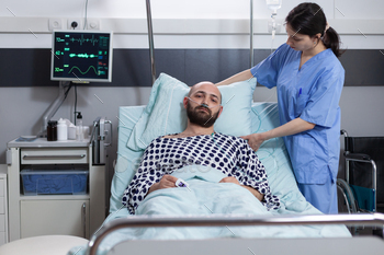 spiratory insufficiency connected to vitals monitor showing low oxygen saturation. Caregiver making man with illness comfortable in hospital bed.