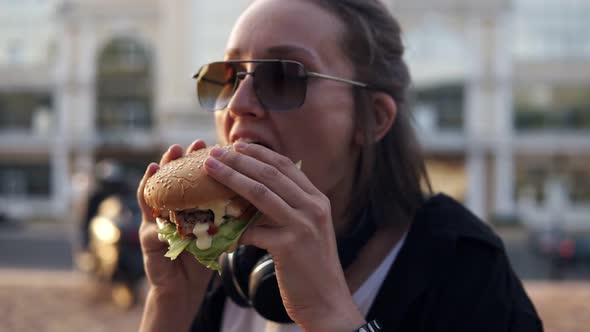 Attractive Young Woman Smiling Cheerfully Holds Tasty Burger in Two Hands and Eats It