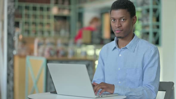 Young African Man with Laptop Smiling at Camera in Cafe