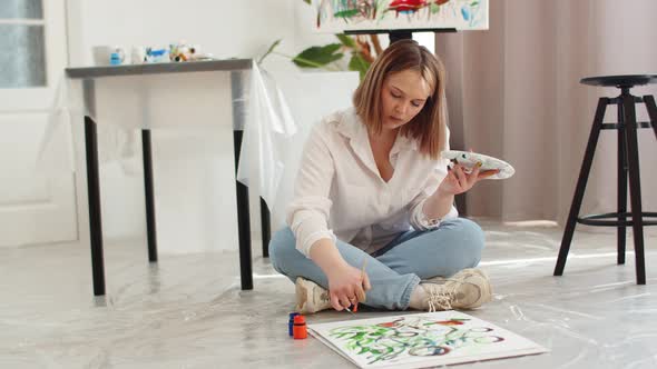 Young Female Artist Paints on Canvas with Brush Dipping It in Paint While Sitting on Floor Front