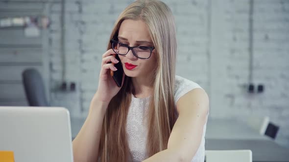 Busy Young Woman Sitting in Office and Speaking on Cellular