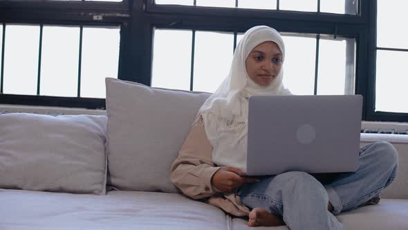 Young Muslim Girl Sits on the Couch Looking at the Laptop in Bright Beige Studio