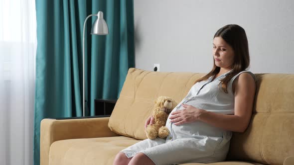 Young Pregnant Woman Stroking Belly Holding Toy Bear While Sitting on Sofa