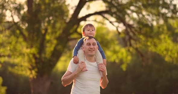 Loving Father Smiles Walking with the Child Sitting on the Neck at Sunset on a Meadow in Summer in