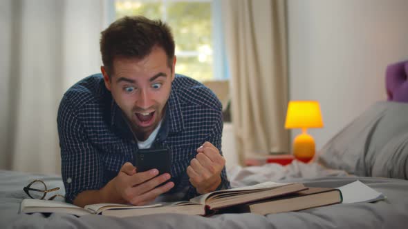 Excited Young Man Student Using Mobile Phone Lying on Bed and Studying