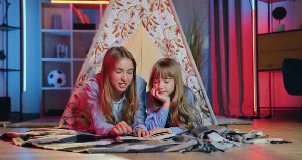two sisters of different ages relaxing on bedcovers in decorative tent and talking about book