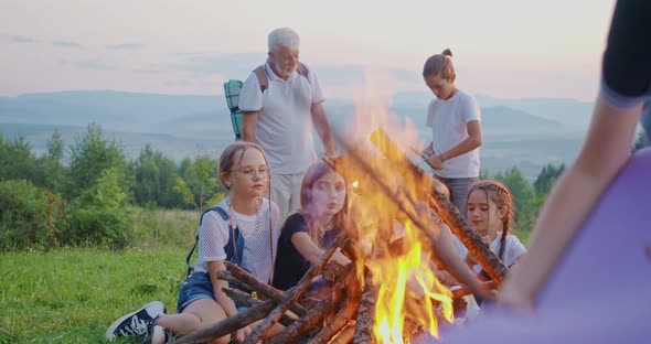 Side View of Kids Sitting on Grass Near Fireplace Old Man with Gray Hair and Rucksack Standing