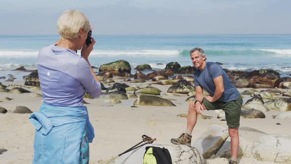 Senior hiker woman taking pictures of senior hiker man using digital camera on the beach.