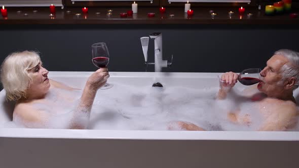 Senior Couple Man and Woman Lying in Warm Bath with Bubbles, Enjoying Relaxation, Drinking Red Wine