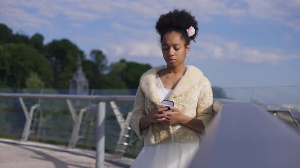 Medium Shot Portrait of Sad Unwilling Bride in Wedding Dress Standing on Bridge Holding Rings Box