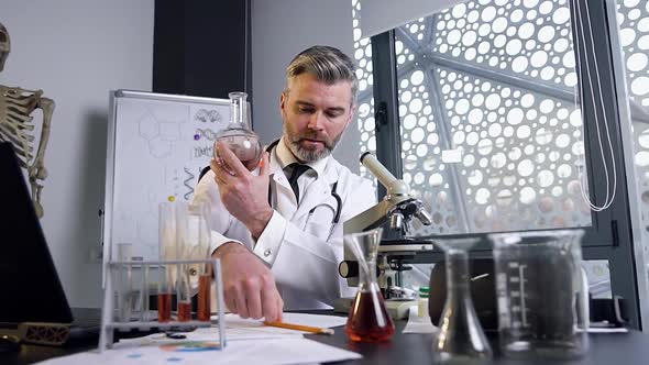 Middle-Aged Scientist Examines the Chemical Liquid in the Flask Sitting at the Table in Lab